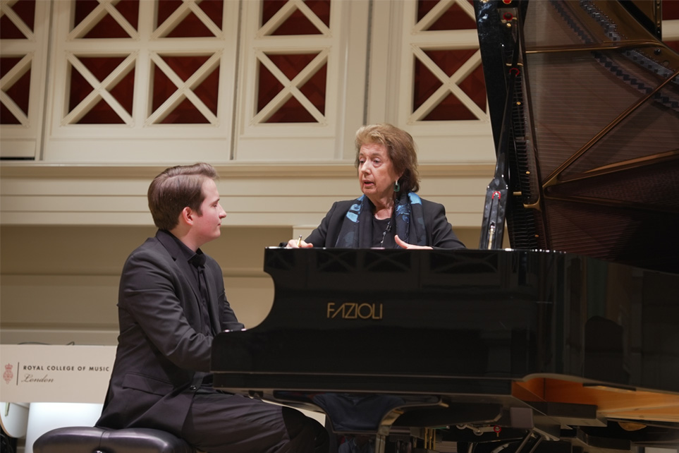 A women wearing a smart blouse with short brown hair, leaning on a piano, talking to a student, wearing smart black attire, looking and listening to her as he sits at the piano, standing on stage.